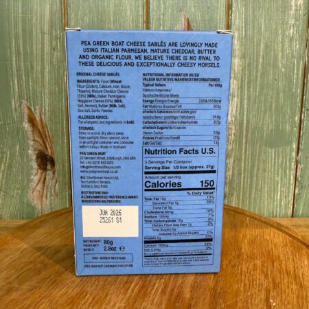 The back of a blue Pea Green Boat Cheese Sables, Original box shows ingredients, nutrition facts, and a June 2026 best-before date, set against a green wooden background decorated with feathers.