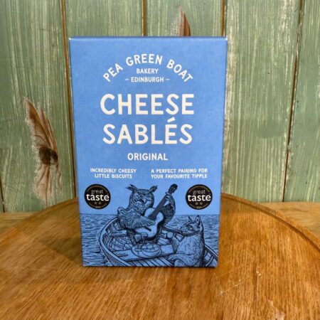 A blue box of Pea Green Boat Cheese Sables, Original rests on wood before a green wall, featuring an illustration of animals in a boat and text calling the biscuits “incredibly cheesy.”.