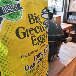A close-up of a yellow bag labeled “Big Green Egg charcoal Large” beside a green ceramic Big Green Egg grill on a wooden table indoors.
