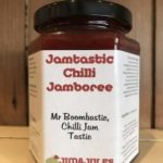 A jar of Jamtastic Chilli Jamboree by Jim & Jules Big Adventure sits on a wooden surface, featuring the text “Mr Boombastic, Chilli Jam Tastie” on its label.