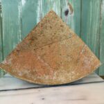 A wedge of textured, tan-colored cheese stands upright on a marble surface, with a green wooden wall in the background.