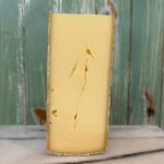 A wedge of yellow cheese with a natural rind and visible cracks sits upright on a marble surface. The background consists of green wooden boards with faint brown spots.