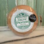 A round wheel of Celtic Promise farmhouse cheese, wrapped in plastic, sits on a marble surface with a wooden background. The label highlights it as washed-rind raw milk cheese and displays a "Great Taste 2023" award.