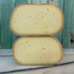 Two halves of a round cheese wheel with a pale yellow interior and natural rind are stacked on a marble surface, with a rustic teal wooden background behind them.