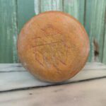 A round loaf of bread with a crisscross pattern on the bottom crust, resting on a marble surface in front of a green wooden background.