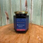 A hexagonal glass jar of Dundarave Blackcurrant & Lime jam sits on a wooden surface in front of a green wooden panel background. The jar has a gold lid and a dark blue label with decorative floral designs.