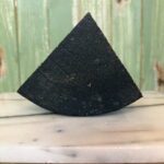 A wedge-shaped piece of black cheese sits on a white marble surface with a green wooden background.