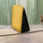 A wedge of hard yellow cheese with a textured black rind sits on a marble surface, with a green wooden background behind it.