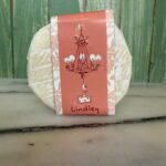 A round wheel of cheese with a pink label showing a chandelier and a small cheese illustration, labeled "Lindley," sits on a marble surface with a green wooden background.