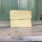 Two halves of a creamy cheese with a white rind are stacked on a marble surface in front of a green wooden wall. The inside of the cheese appears smooth and pale yellow.