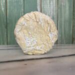 A round wheel of soft, white-rinded cheese sits on a marble surface with a green wooden background.