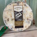 A wheel of Quicke's Goat's Milk Clothbound Cheese with a white and marbled rind, wrapped in cloth, sits on a white surface with a green wood background. The label features a black illustration and gold accents.