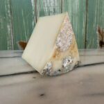 A wedge of cheese with a hard, rind-coated exterior showing blue and white mold spots, sitting on a marble surface in front of a green wooden background.