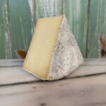 A wedge of hard cheese with a rough, grayish rind and pale yellow interior sits on a marble surface in front of a green wooden background.