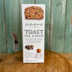 A box of The Fine Cheese Co. Toast for Cheese, featuring apricots, pistachios, and sesame seeds, is displayed on a wooden surface with a green wooden background.