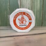 A wheel of Village Maid Waterloo Guernsey milk cheese in white wrapping with an orange label showing an illustration of people around a maypole, set against a green wooden background and a marble surface.