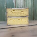Two wedges of soft, creamy cheese with a white rind and small holes, stacked on a marble surface. The background features vertical green wooden panels.