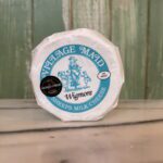A round package of Village Maid Wigmore sheep’s milk cheese sits on a marble surface, with a green wooden wall in the background. The label features a blue illustration and a black World Cheese Awards sticker.