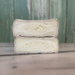 Two halves of a creamy, white cheese with a soft rind stacked on a marble surface in front of a green wooden background.