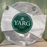A large round wheel of Cornish Yarg cheese wrapped in paper labeled "YARG" and "Nettle Covered", with a dark green circle at the center displaying the cheese’s name and description. The wheel sits on a marble slab.
