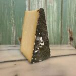 A wedge of cheese with a dark, rustic rind and firm pale yellow interior sits upright on a marble surface in front of a green wooden wall.