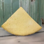 A wedge of yellow cheese with a firm texture sits on a marble surface in front of a green wooden background.