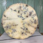 A close-up of a wheel of blue cheese, showing its creamy texture and blue-green veins, set against a green wooden background and resting on a marble surface.