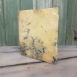A wedge of blue cheese with blue veins running through it, placed on a marble surface in front of a green wooden background.