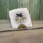 A round package of goat cheese labeled "URAFFR" sits on a marble surface, in front of a green wooden wall. The label features an illustrated goat’s head with horns and cheese award emblem.