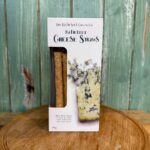 A box of Bath Blue Cheese Straws by The Bath Soft Cheese Co. is displayed on a round wooden board with a green wooden background. The packaging shows cheese straws and a piece of blue cheese.