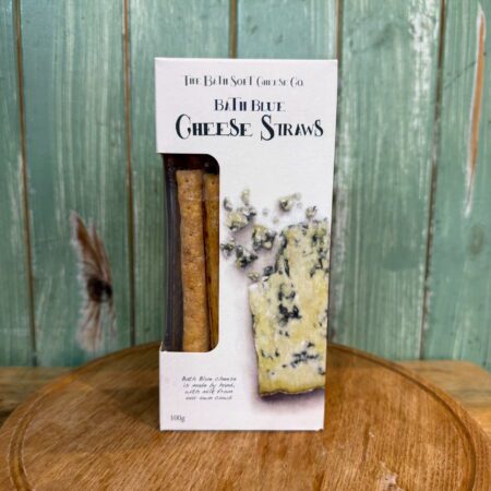 A box of Bath Blue Cheese Straws by The Bath Soft Cheese Co. is displayed on a round wooden board with a green wooden background. The packaging shows cheese straws and a piece of blue cheese.