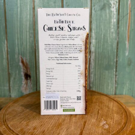 A box of Bath Blue Cheese Straws stands on a wooden surface against a green wooden plank background. The box details ingredients, nutritional information, allergens, and company details in black text.