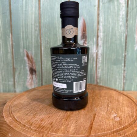 A dark glass bottle of extra virgin olive oil with a black cap sits on a round wooden board. The bottle’s label, featuring product information and a QR code, faces the camera. The background is wooden paneling.