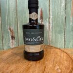 A bottle of Two & One extra virgin olive oil sits on a wooden surface with a green wooden background. The label notes it is a product of Spain, cold extracted, with premium pressing and early harvest.