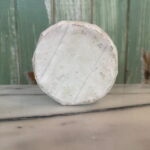 A round wheel of brie cheese with a white rind sits upright on a marble surface, with a rustic green wooden wall in the background.