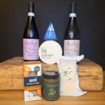 The Christmas Hamper £65 (Copy) features two bottles of wine, blue cheese, Baron Bigod, hard cheese, sourdough crackers, and rhubarb apple & fig chutney arranged on a wooden surface against a black background.
