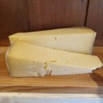 Two thick wedges of Montgomery Cheddar 250g, with a crumbly texture and yellow hue, are placed on a wooden board in front of a wooden wall background.