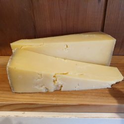 Two thick wedges of Montgomery Cheddar 250g, with a crumbly texture and yellow hue, are placed on a wooden board in front of a wooden wall background.