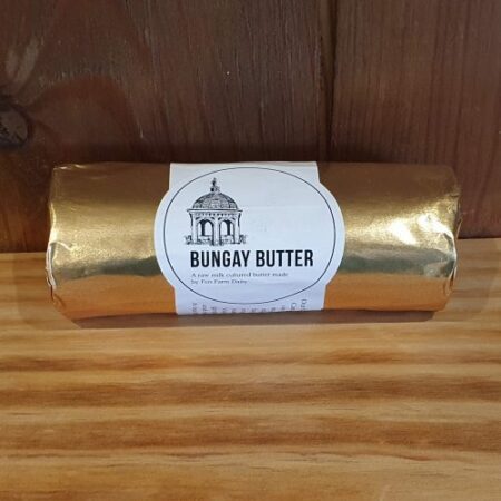 A roll of Bungay Butter wrapped in gold foil with a white label featuring a building illustration, set on a wooden surface against a wooden background.