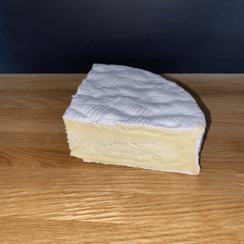 A 250g wedge of Baron Bigod cheese, featuring a soft, creamy texture and white rippled rind, rests on a wooden surface with a dark backdrop.