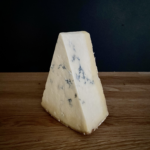 A 250g wedge of Cropwell Bishop Organic Stilton blue cheese with visible veins rests on a wooden surface against a dark background.